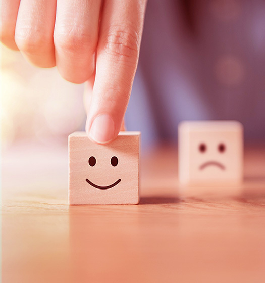 A hand holds up a wooden tile with a smiley face printed on it on a table. In the background, out of focus, is a tile with a sad face.