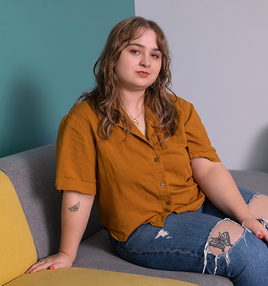 A young woman sits on a couch looking into the camera. Tattoos are visible through the tears in the knees of her blue jeans.